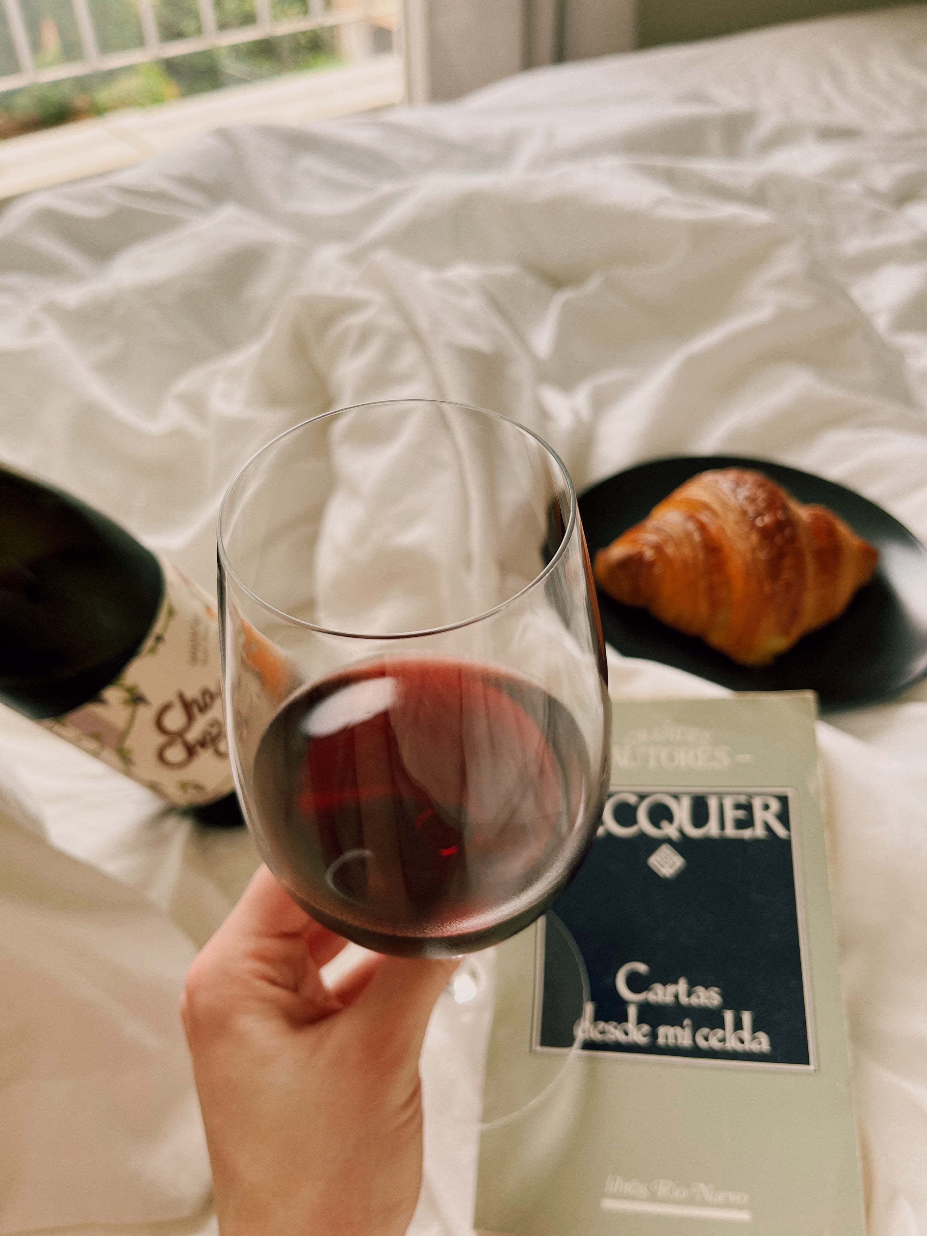 Glass of Cha Cha Cha red wine held by a hand on a bed with a book and pastry.
